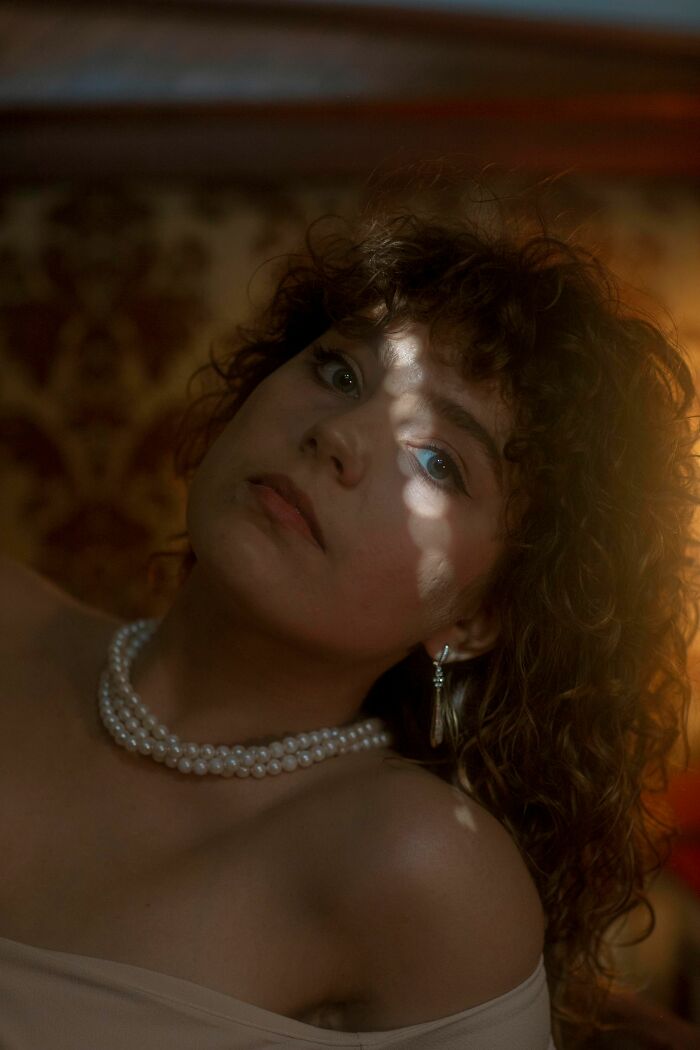 Woman with curly hair and pearl necklace looking up, with light hitting her face, suggesting sick burns of emotion.