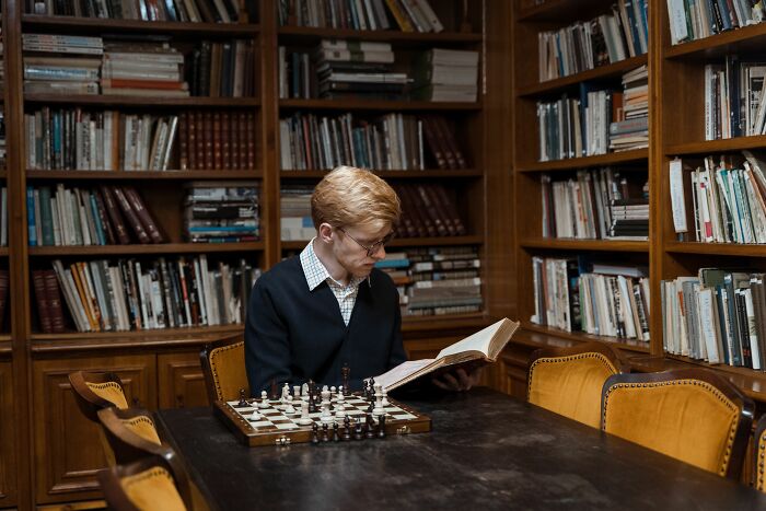 A man with red hair and glasses in a library, reading a book. A chessboard is on the table, showcasing high IQs and general knowledge questions. A man with red hair and glasses in a library, reading a book. A chessboard is on the table, showcasing high IQs and general knowledge questions.