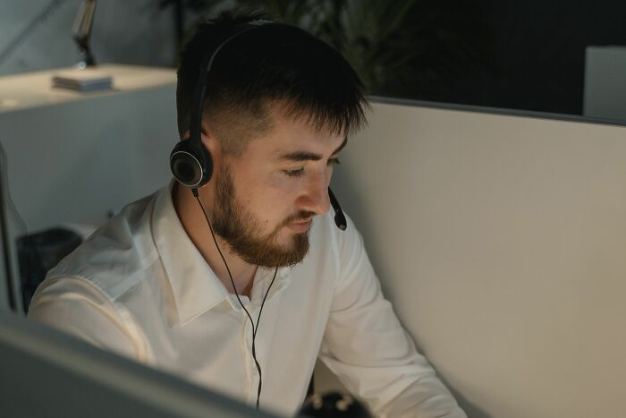 A man with a beard wearing a headset and working at a desk, looking tired, reflecting office work drama stories.