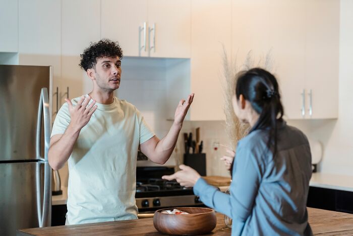 A man with curly hair gestures with his hands while talking to his girlfriend in a kitchen setting.