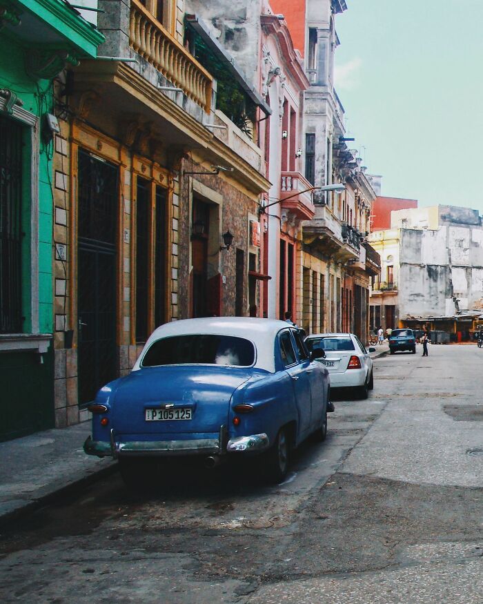 A vintage blue car parked on a narrow street in a city, with colorful buildings lining the road. People believe today that propaganda influences beliefs.