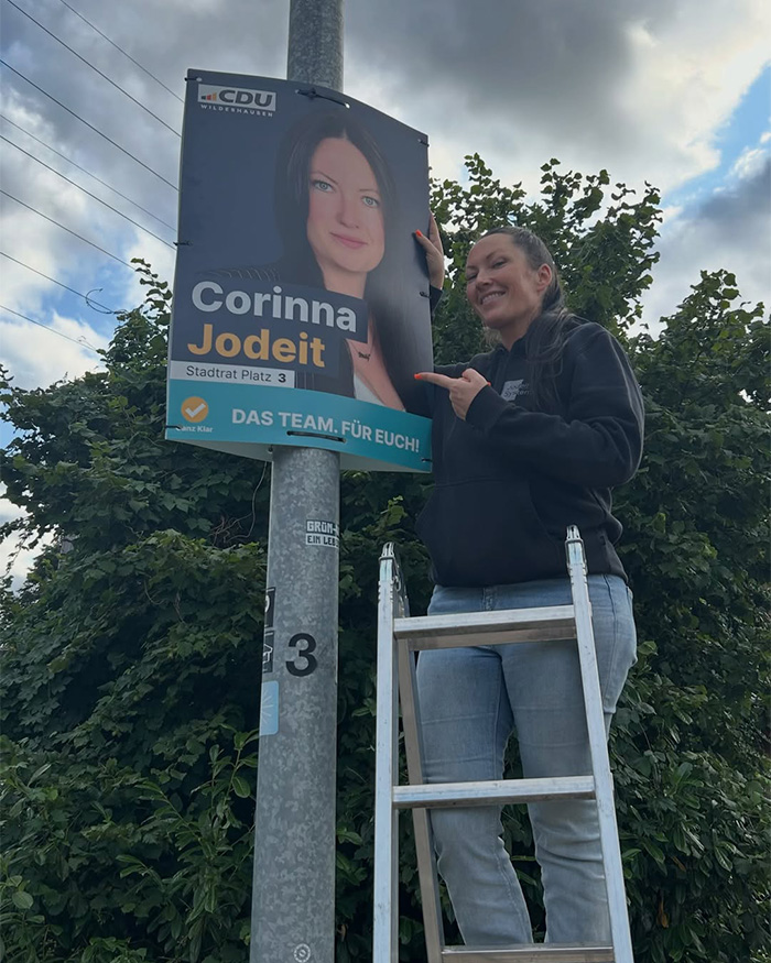 Conservative politician Corinna Jodeit on a ladder, pointing at her campaign poster with a provocative necklace. Conservative politician Corinna Jodeit on a ladder, pointing at her campaign poster with a provocative necklace.