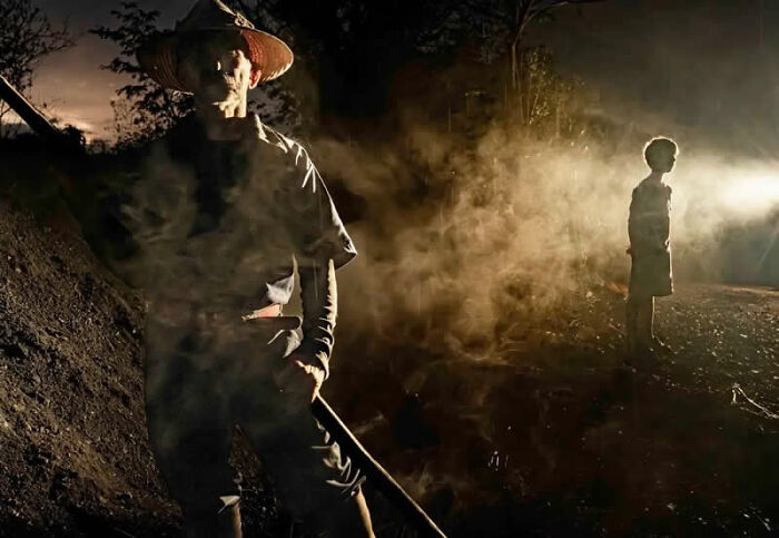 A person in a straw hat stands in a smoky, dark setting, a street photo capturing life's coincidences.