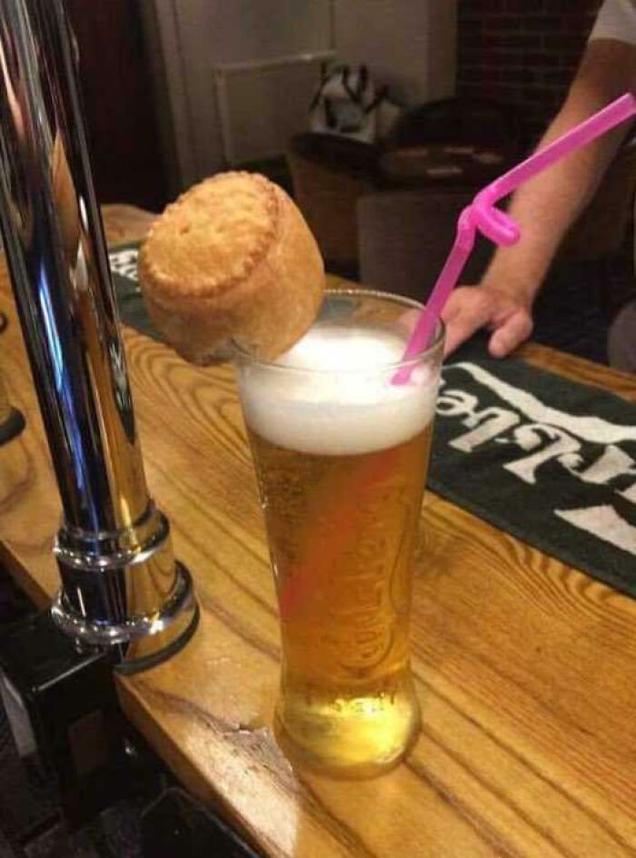 A pint of beer with a small savory pie perched on its rim and a pink straw, showcasing British humor.