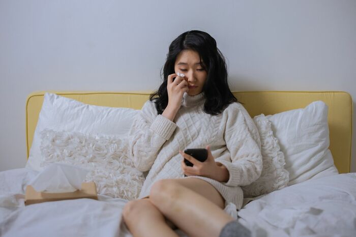 A young woman crying and wiping her eye while looking at her phone, with a tissue box next to her.
