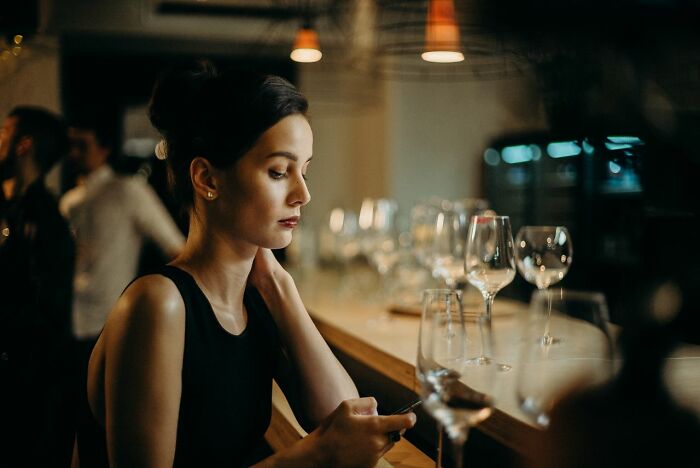 A woman sitting alone at a bar, looking at her phone, depicting the sadness when friendships fell apart.