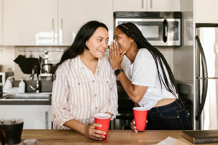Two friends in a kitchen, one whispering to the other, portraying the complexities of friendships that fell apart.
