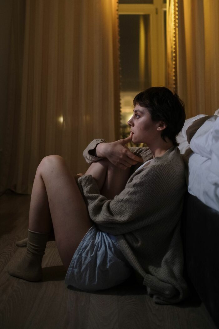 A young woman with short hair sitting on the floor in a dim room, looking pensive.