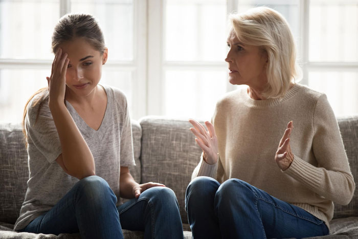 A young bride looks upset while her mother gesticulates angrily on a couch, discussing the aisle walk.