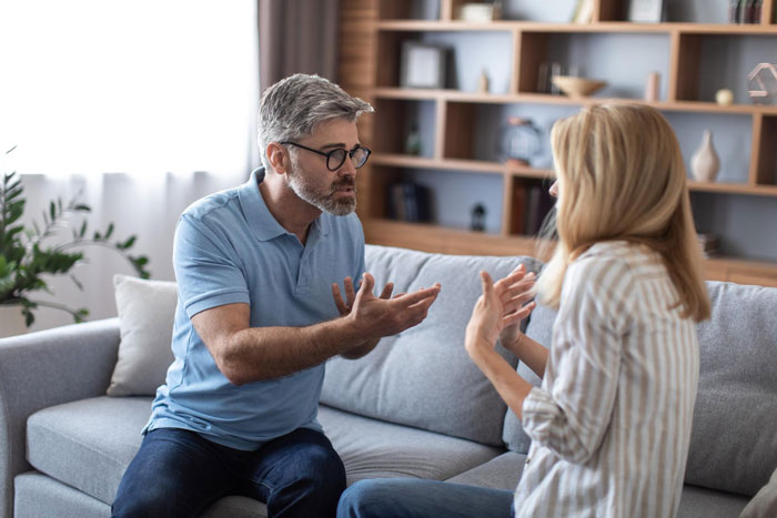 A man and woman, possibly a dad and mom, sit on a couch engaged in a serious discussion about the bride walking down the aisle.