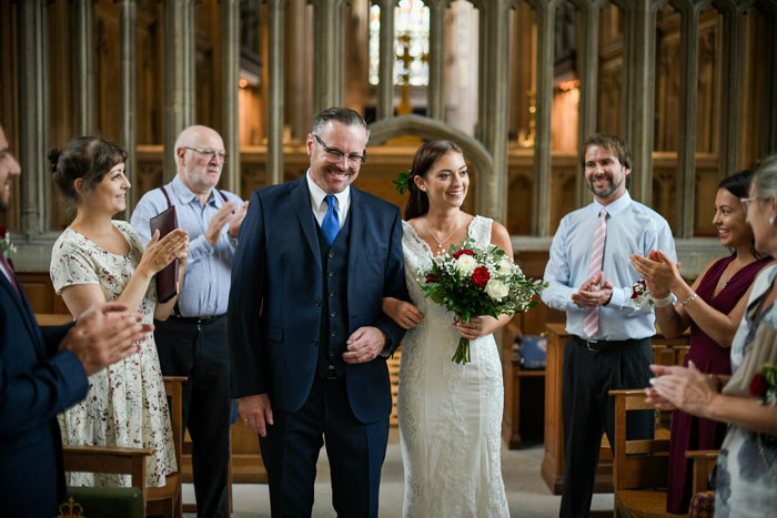 A beaming bride in a white lace gown with a red and white bouquet walks down the aisle with her dad during her wedding ceremony, surrounded by applauding guests.