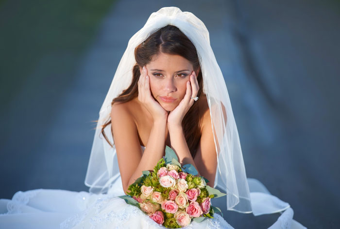 A thoughtful bride, wearing a wedding dress and veil, sits with a bouquet, contemplating who should walk her down the aisle.