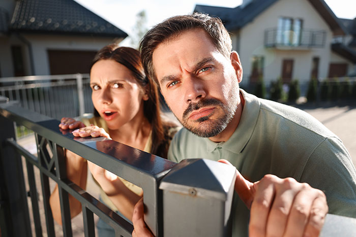 Man and woman peeking over a fence, looking concerned about HOA rules and their home and land situation. Man and woman peeking over a fence, looking concerned about HOA rules and their home and land situation.