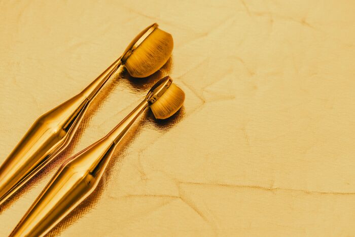 Two golden makeup brushes on a shiny golden surface, representing the luxurious items owned by the rich.