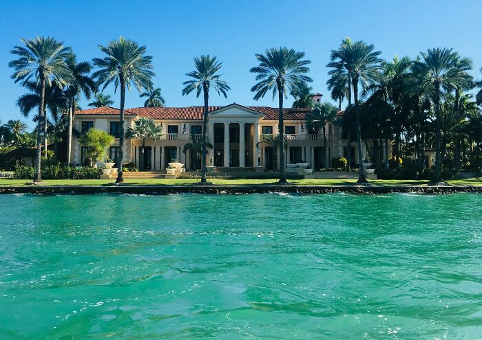 A grand mansion on the water, surrounded by palm trees, showcasing the lavish lifestyle of the rich.