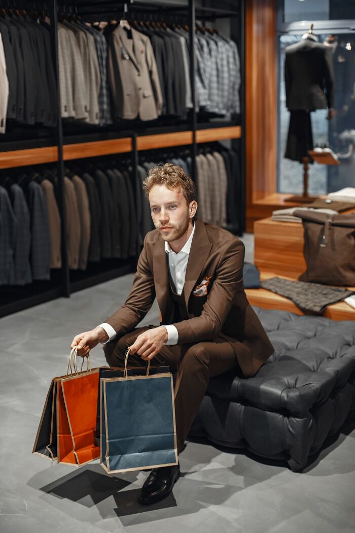 A man in a brown suit sits in a high-end clothing store with shopping bags, depicting wild moments for the rich.