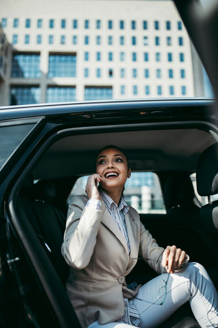 A smiling businesswoman in a suit talks on her phone in the back of a luxury car, reflecting moments of the rich.
