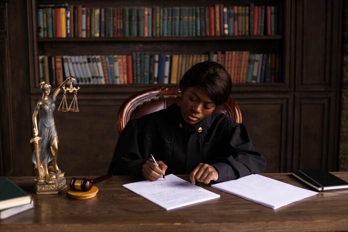 A judge in robes writing at a desk with a Lady Justice statue and gavel. This image is central to court case stories.