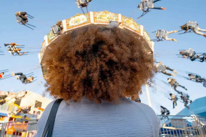 A person with curly hair views a spinning carousel ride with people flying high, capturing life's funniest coincidences in street photos.