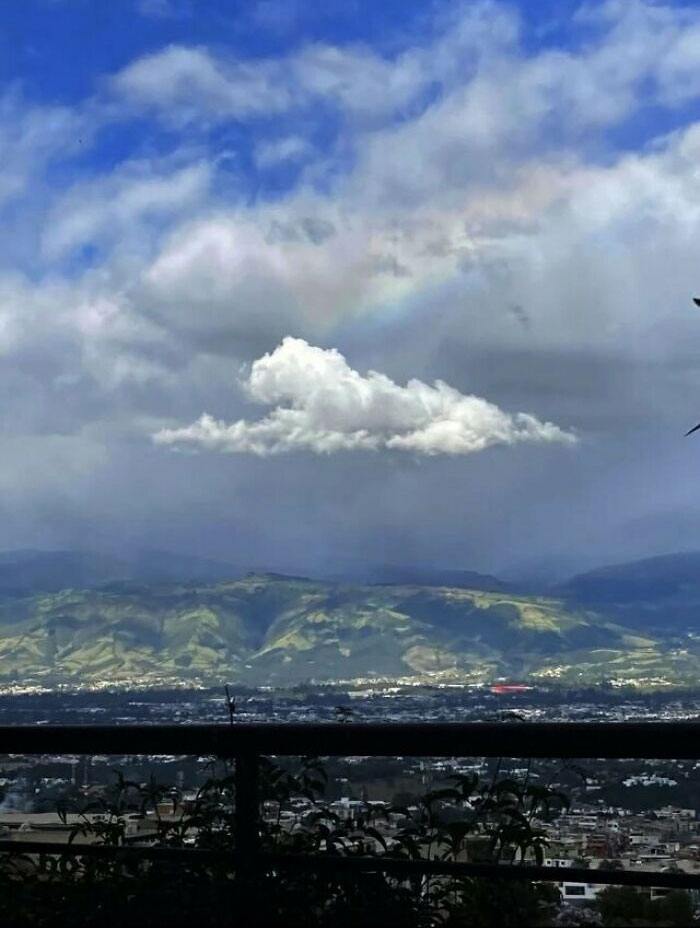 A rainbow over a single cloud in a blue sky above mountains and a city, a sign from the universe.