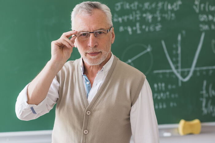 An older male teacher in a classroom, standing in front of a chalkboard with equations, a wise sign from the universe.