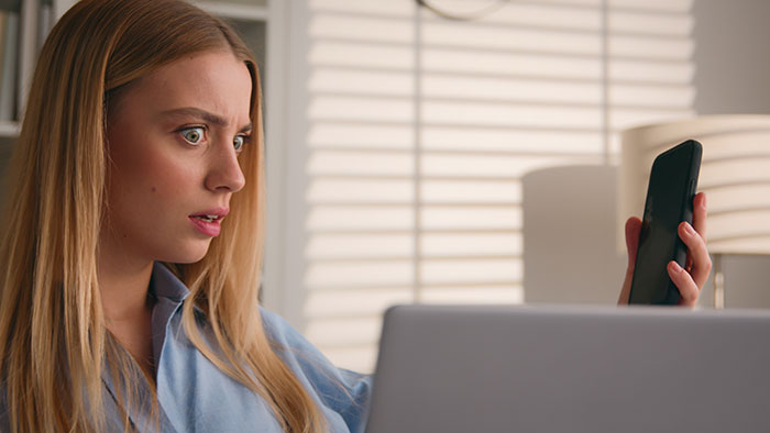 A woman with tech help from a coworker looking shocked while holding her phone and looking at her laptop.