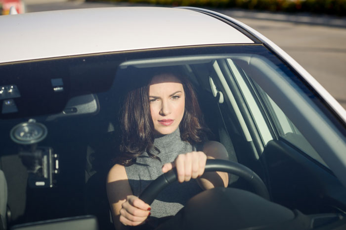 A woman with dark hair driving a white car, looking forward, relevant to the SIL bullying and hearing aids. A woman with dark hair driving a white car, looking forward, relevant to the SIL bullying and hearing aids.