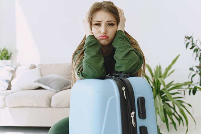 A sad woman with long brown hair, wearing a green outfit, sitting behind a blue suitcase, upset about her half-sis and infertility.