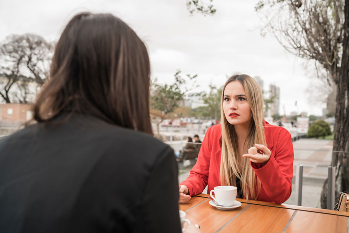 Two women having a serious conversation at an outdoor cafe. One woman in red gestures, discussing infertility.