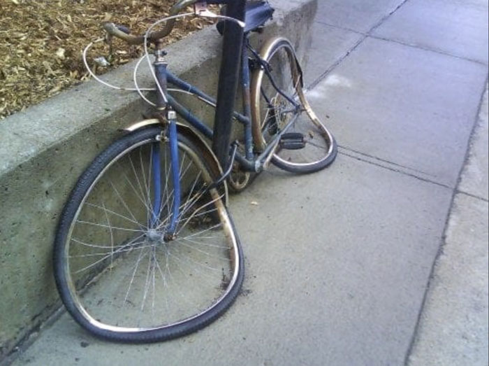 A bicycle with both wheels completely warped and melted, leaning against a concrete curb due to summer heat.