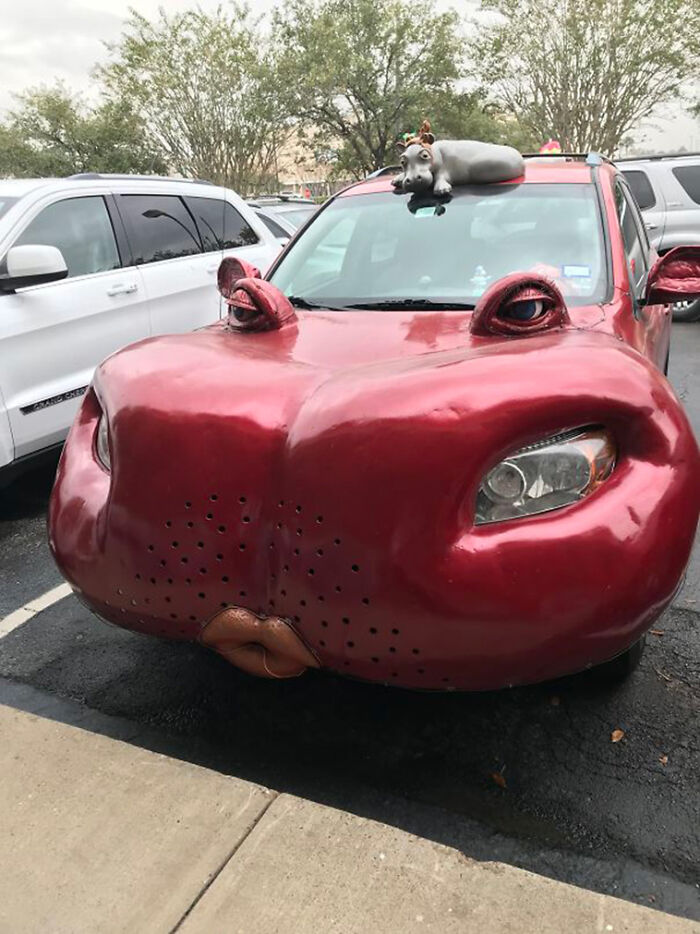 A red car with a giant hippo head and a small hippo on the roof, an awful idea executed with skill.