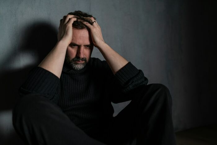 A man with a beard sits on the floor, holding his head in his hands, depicting the struggle with mental health disorders.