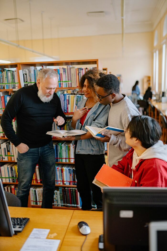 An older man with a beard helping young students in a library, exemplifying things America does better.