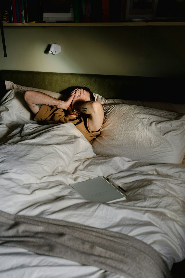 A person in bed, hands over their face, a notebook nearby, showing interesting neighbors.
