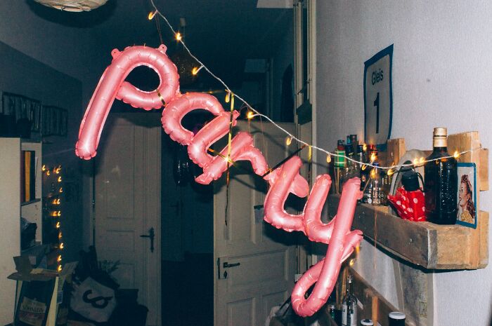 Pink balloons spelling out PARTY with string lights, hanging in a dimly lit hallway. The scene suggests interesting neighbors celebrating.
