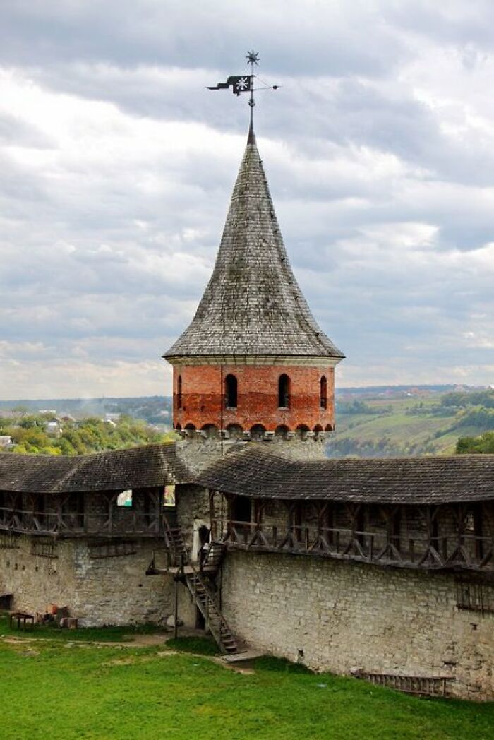 An ancient stone castle tower with character, featuring a conical roof and a weathered brick upper section under a cloudy sky.