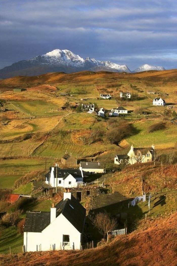 A cluster of picturesque white buildings with character nestled in a rolling green landscape, with snow-capped mountains.