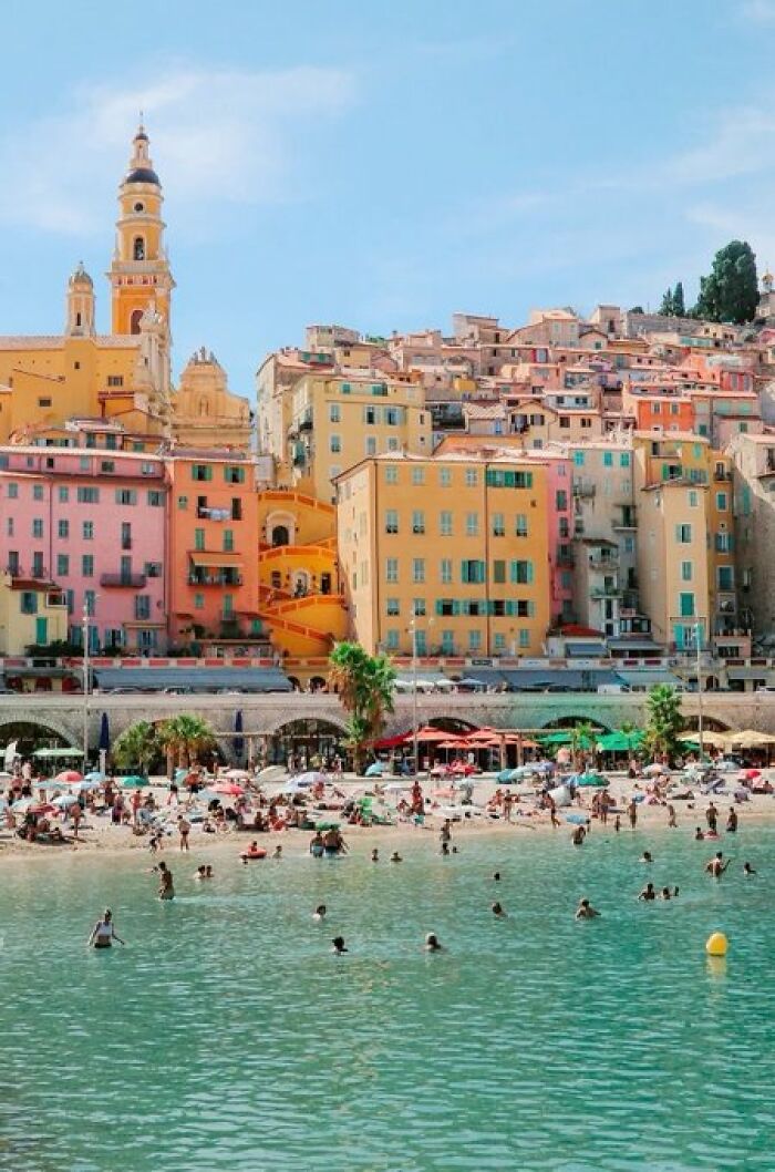A colorful coastal town with buildings of unique character overlooks a bustling beach and vibrant blue sea.