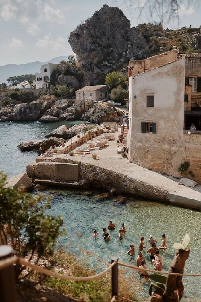 Swimmers enjoy clear turquoise waters next to a rocky shoreline village, featuring buildings with old-world character.