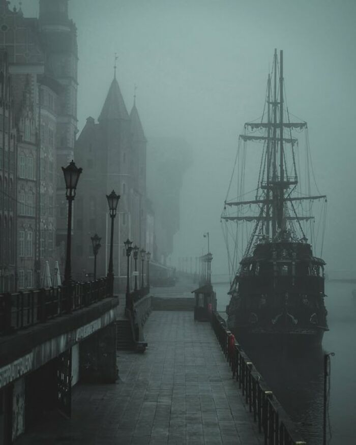 A misty harbor scene with an old sailing ship docked next to historic buildings with character.