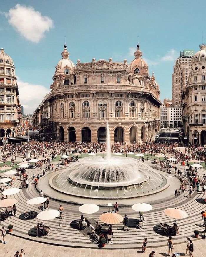 Grand, historic building with character and a large fountain in a bustling city square, surrounded by people and umbrellas.