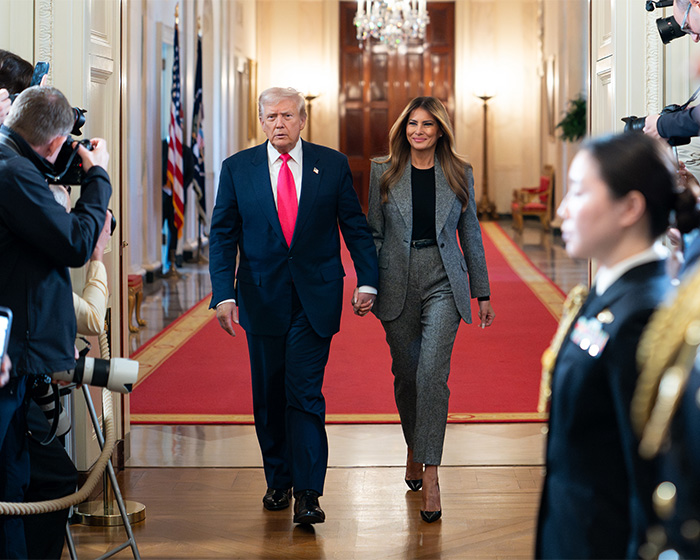Donald Trump and Melania in formal attire, walking down a red-carpeted hallway, facing media scrutiny.