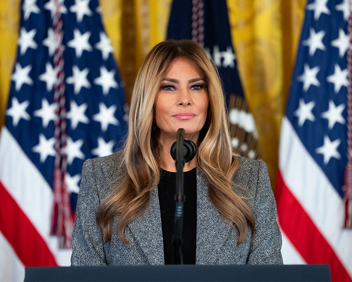 Melania Trump at a podium, speaking, with US flags in the background, amid public scrutiny.