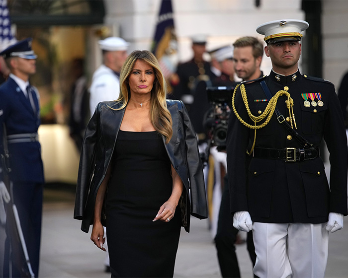 Melania in a black dress and leather jacket walking next to a military officer, with other officers in the background. Melania's absence.