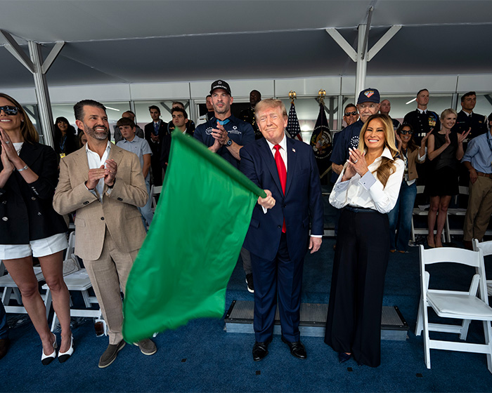 Donald Trump holding a green flag, flanked by Melania Trump and others, as Natalie Harp appears in the scene. Donald Trump holding a green flag, flanked by Melania Trump and others, as Natalie Harp appears in the scene.