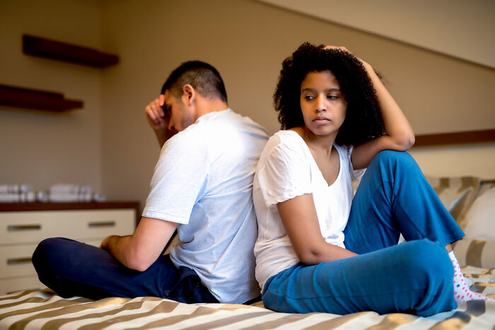 A diverse couple sitting back to back on a bed, looking upset, considering divorce papers and fighting harder.