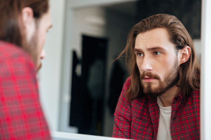 A man with long hair and a beard looking intently into a mirror, contemplating why he signed divorce papers.
