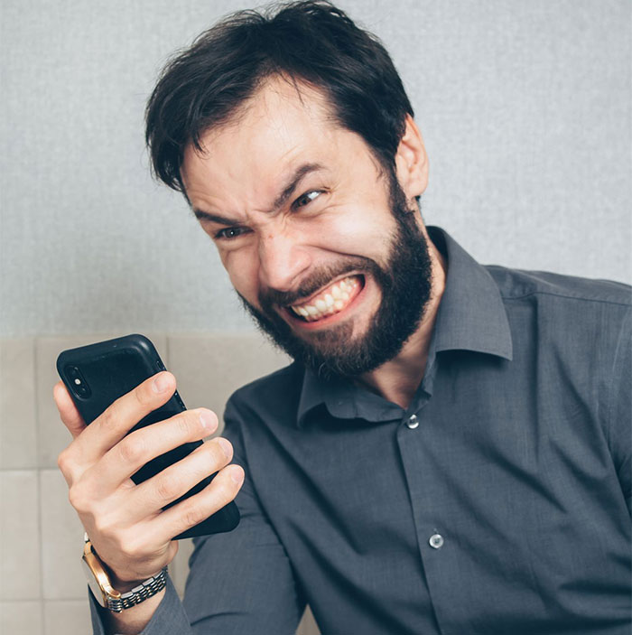 A man with a beard and dark shirt, looking angry and grimacing while holding a smartphone, representing a bully. A man with a beard and dark shirt, looking angry and grimacing while holding a smartphone, representing a bully.
