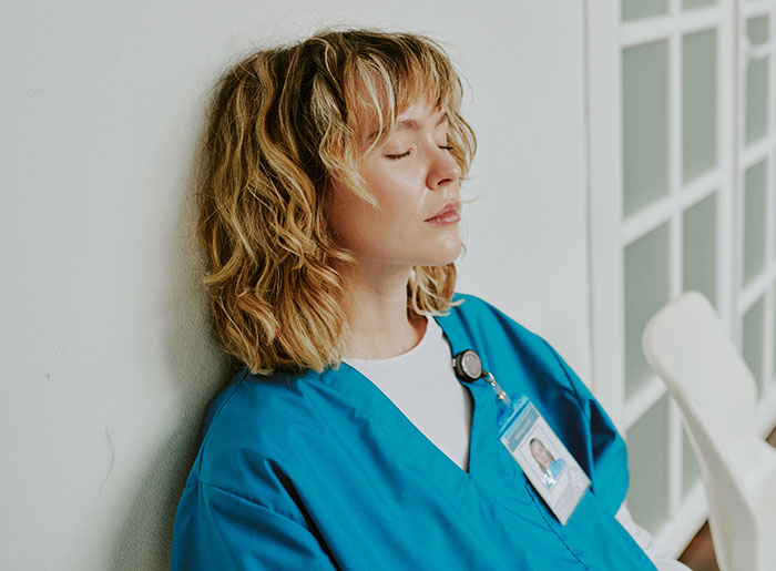 A nurse with closed eyes, leaning against a wall, appearing tired, after patients said weird things.