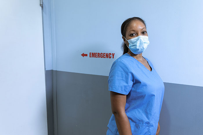 A nurse wearing a mask in a hospital hallway, near an emergency sign, listening to patients saying weird things.